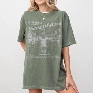 以下が含まれることがあります： オリーブグリーンのTシャツ。ディズニーランドのグラフィックがプリントされています。グラフィックにはミッキーマウス、「Disneyland」と「California Est. 1955」の文字、「The happiest place on earth」のフレーズが含まれています。
