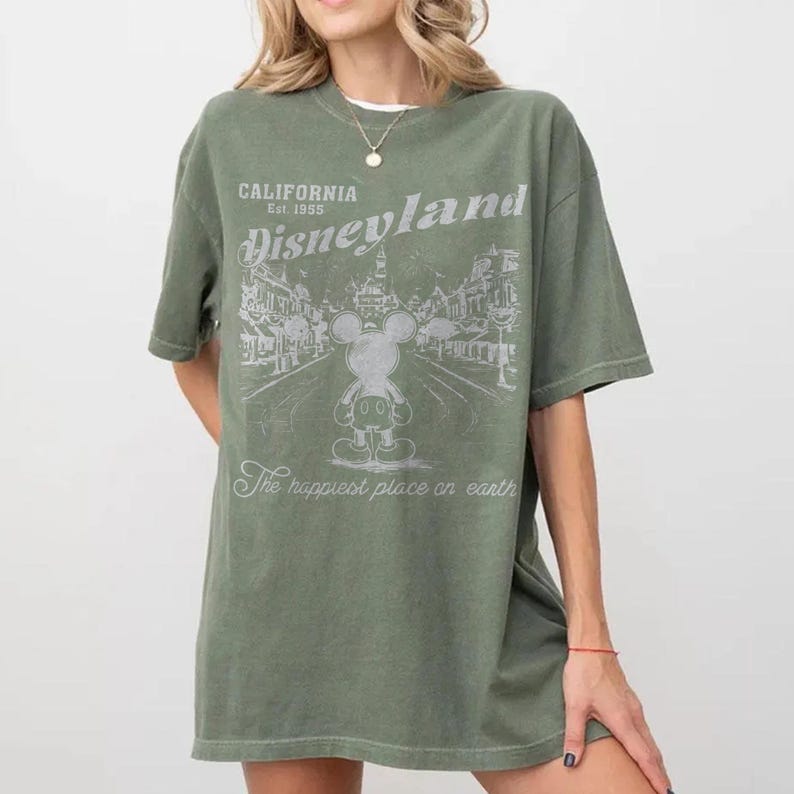 以下が含まれることがあります： オリーブグリーンのTシャツ。ディズニーランドのグラフィックがプリントされています。グラフィックにはミッキーマウス、「Disneyland」と「California Est. 1955」の文字、「The happiest place on earth」のフレーズが含まれています。