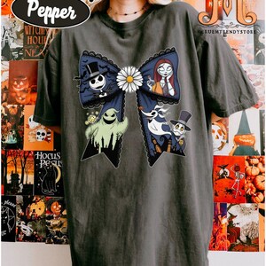 Peut inclure: T-shirt gris fonc&eacute; avec un grand motif de n&oelig;ud orn&eacute; de personnages de L'&Eacute;trange No&euml;l de Monsieur Jack. Le n&oelig;ud bleu marine est bord&eacute; de dentelle noire et orn&eacute; d'une marguerite blanche. Les personnages incluent Jack Skellington, Sally, Zero et Oogie Boogie.