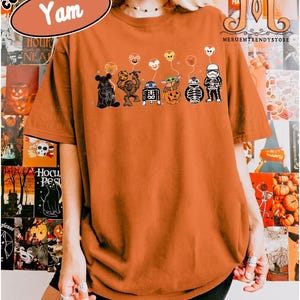 Op de afbeelding: Een Yam-kleurig Comfort Colors t-shirt met een Halloween-thema. De afbeelding toont stripfiguren met pompoenelementen en ballonnen. Het shirt heeft de tekst "Comfort Colors" en "Yam" linksboven.