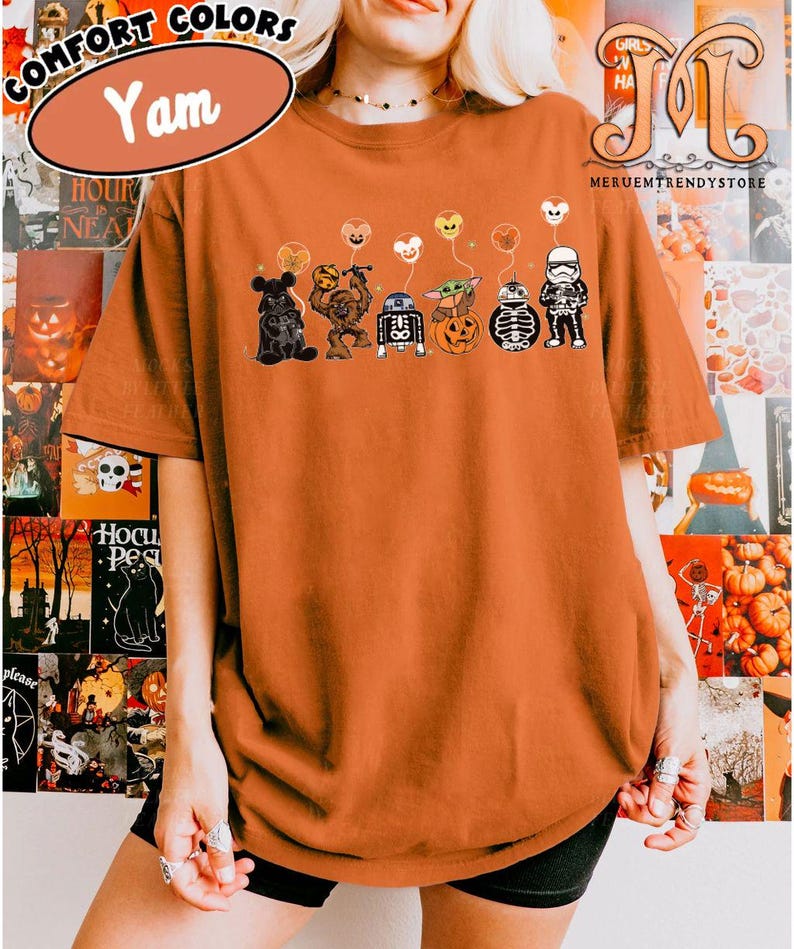 Op de afbeelding: Een Yam-kleurig Comfort Colors t-shirt met een Halloween-thema. De afbeelding toont stripfiguren met pompoenelementen en ballonnen. Het shirt heeft de tekst "Comfort Colors" en "Yam" linksboven.