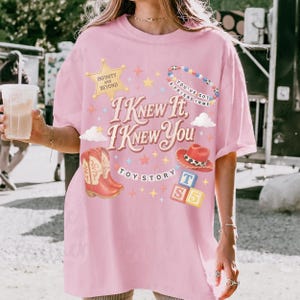 May include: A light pink t-shirt featuring a graphic print with the text "I KNEW IT, I KNEW YOU", "INFINITY AND BEYOND", "YOU'VE GOT A FRIEND IN ME", "TOY STORY", and "T S 5". The design includes a gold star, cowboy boots, a hat, and blocks.