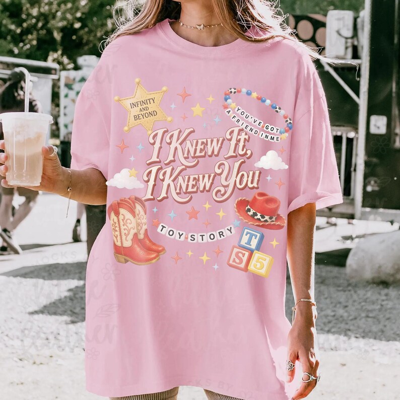 May include: A light pink t-shirt featuring a graphic print with the text "I KNEW IT, I KNEW YOU", "INFINITY AND BEYOND", "YOU'VE GOT A FRIEND IN ME", "TOY STORY", and "T S 5". The design includes a gold star, cowboy boots, a hat, and blocks.
