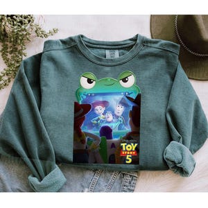May include: Teal crew neck sweatshirt with a frog-shaped frame displaying characters from Toy Story 5. The sweatshirt has rolled cuffs and a relaxed fit. The Toy Story 5 logo is visible at the bottom of the screen.