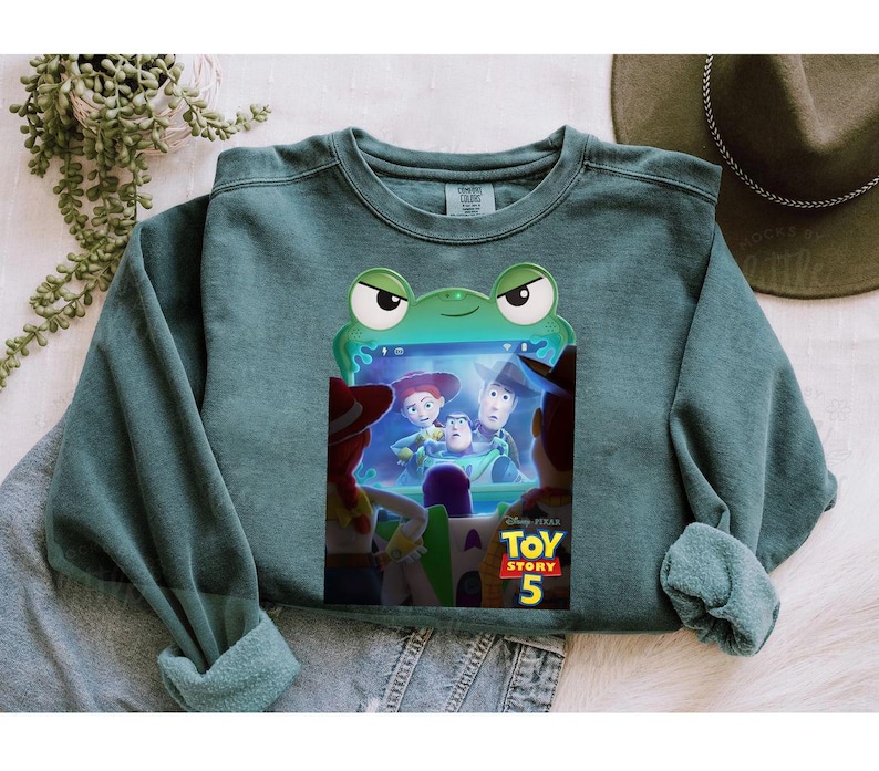 May include: Teal crew neck sweatshirt with a frog-shaped frame displaying characters from Toy Story 5. The sweatshirt has rolled cuffs and a relaxed fit. The Toy Story 5 logo is visible at the bottom of the screen.