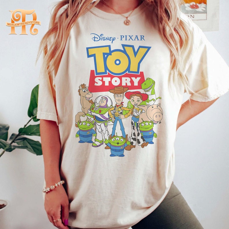 May include: Cream-colored t-shirt featuring the Disney Pixar Toy Story logo and character illustrations. The design includes Woody, Buzz Lightyear, and other characters from the animated film. The shirt has a relaxed fit.