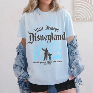 Puede incluir: Camiseta azul claro con el texto "Walt Disney's Disneyland" en una fuente estilizada. Debajo del texto, una silueta de una persona y Mickey Mouse frente a un castillo. La camiseta tambi&eacute;n incluye el texto "The Happiest Place On Earth EST. 1955."
