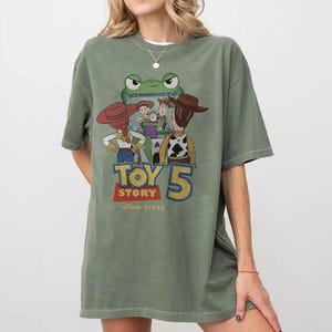 May include: Olive green t-shirt featuring a graphic from the movie Toy Story 5. The graphic includes the characters Woody, Buzz Lightyear, Jessie, and others. The text "TOY STORY 5" is displayed in a colorful font, with the Disney Pixar logo below.