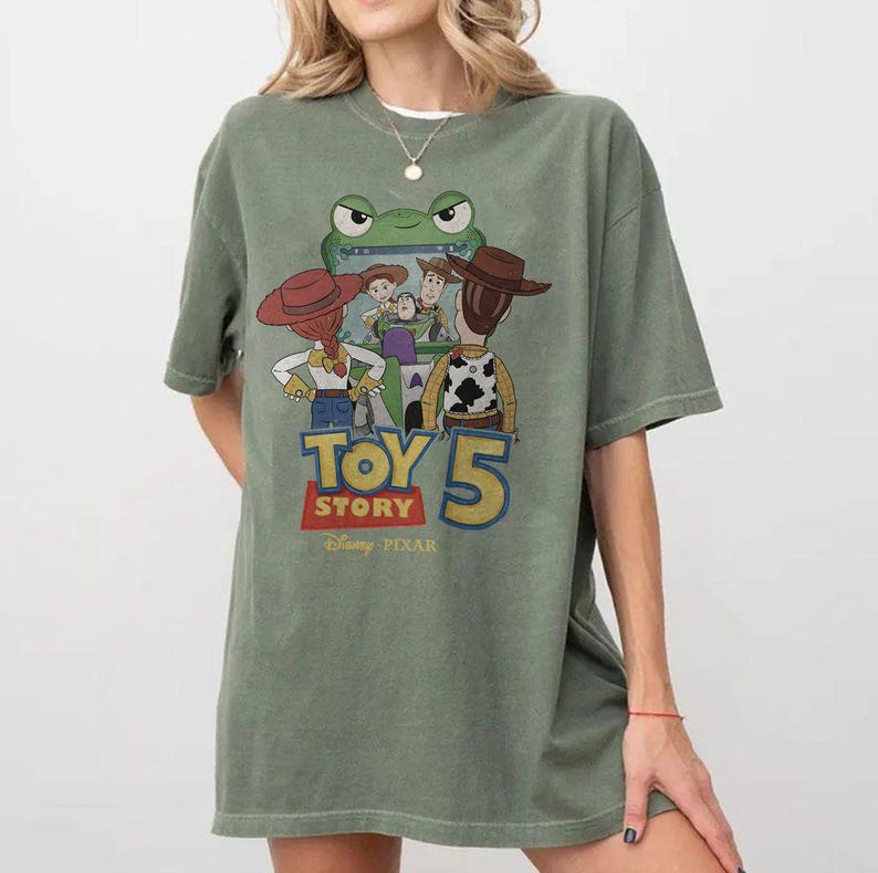 May include: Olive green t-shirt featuring a graphic from the movie Toy Story 5. The graphic includes the characters Woody, Buzz Lightyear, Jessie, and others. The text "TOY STORY 5" is displayed in a colorful font, with the Disney Pixar logo below.