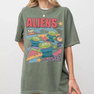May include: Olive green t-shirt featuring the word "ALIENS" in red, with a graphic of three green aliens from Toy Story. The design includes the text "TOY STORY" and "SINCE 1995". The aliens are wearing blue outfits.