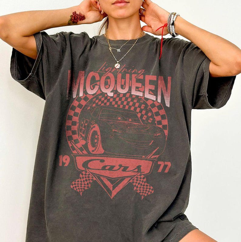 May include: Dark gray Lightning McQueen Cars t-shirt. The graphic features a red and black checkered circle with a car illustration, the text "Lightning McQueen", "Cars", and the numbers "19" and "77". The shirt has short sleeves and a relaxed fit.