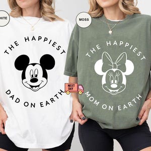 May include: White and moss green t-shirts with Mickey and Minnie Mouse graphics. The white shirt says "The Happiest Dad on Earth" and the green shirt says "The Happiest Mom on Earth". Both shirts have black text and graphics.