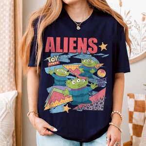 May include: Navy blue t-shirt featuring the word "ALIENS" in red, with "TOY STORY" below. The shirt displays three green aliens with three eyes each, set against a colorful geometric background. The text "SINCE 1995" is also included.