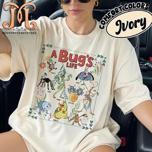 以下が含まれることがあります： 「バグズ・ライフ」のカラフルなグラフィックが特徴のアイボリー色のTシャツ。デザインには、さまざまな漫画の昆虫と「A Bug's Life」の文字が含まれています。シャツには「Comfort Colors」と「Ivory」の文字も入っています。