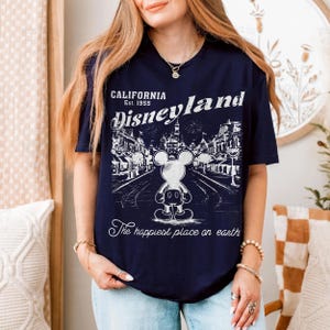 以下が含まれることがあります： ネイビーブルーのTシャツ。白のディズニーランドのグラフィックで、ミッキーマウスと「California Est. 1955 Disneyland The happiest place on earth」の文字が入っています。ヴィンテージスタイルのイラストです。
