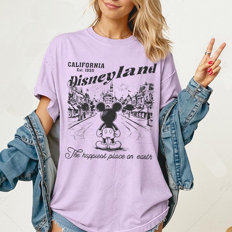 以下が含まれることがあります： ディズニーランドのグラフィックがプリントされた薄紫色のTシャツ。グラフィックには、建物のある通りに立っているミッキーマウスと、「California Disneyland Est. 1955」と「The happiest place on earth」の文字が描かれています。
