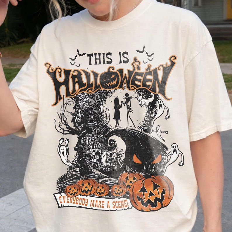 May include: An off-white t-shirt featuring a black and orange graphic print with the text "THIS IS HALLOWEEN" and "EVERYBODY MAKE A SCENE". The design includes silhouettes of characters, ghosts, bats, a large pumpkin, and smaller pumpkins.