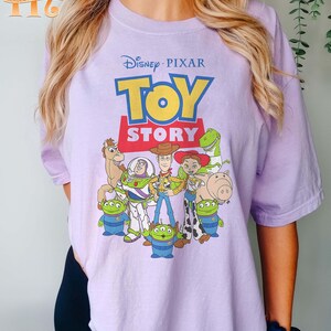 May include: A light purple t-shirt featuring the Disney Pixar Toy Story logo and characters. The graphic includes Woody, Buzz Lightyear, and other characters from the animated film. The shirt has short sleeves and a crew neck.