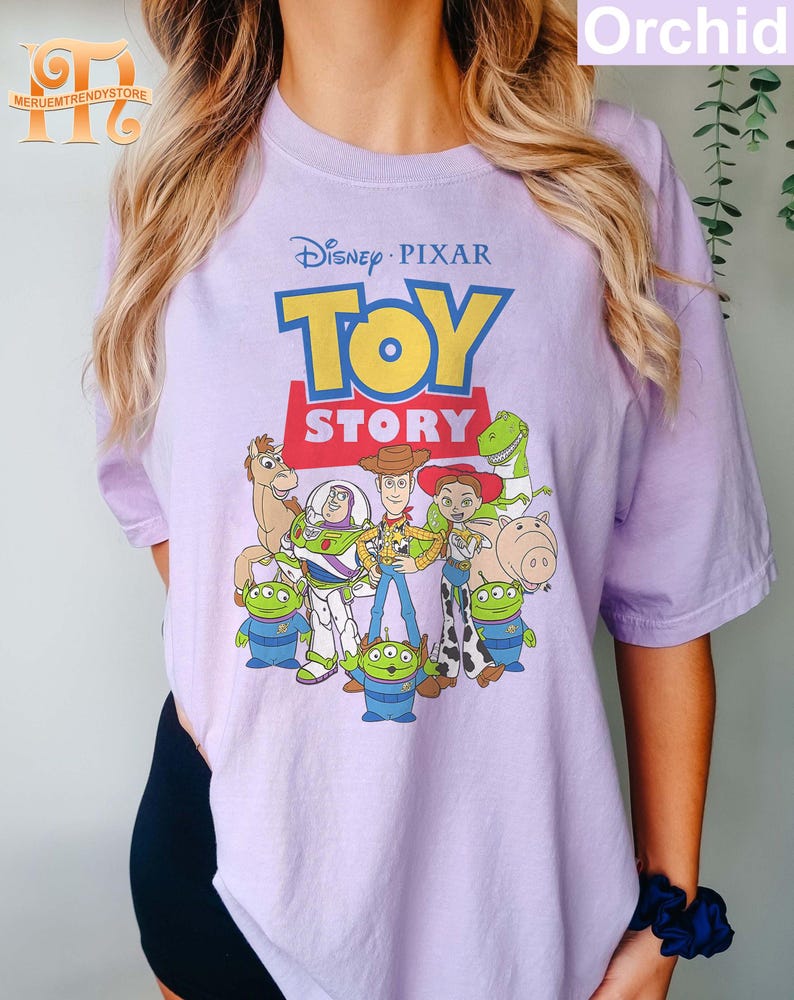 May include: A light purple t-shirt featuring the Disney Pixar Toy Story logo and characters. The graphic includes Woody, Buzz Lightyear, and other characters from the animated film. The shirt has short sleeves and a crew neck.
