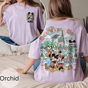 May include: Orchid-coloured t-shirt with a Mickey Mouse graphic and the word "Daddy" on the front. The back features a "Walt Disney World" design with Mickey, Minnie, Donald, Goofy, and Pluto. The shirt is a light purple colour.