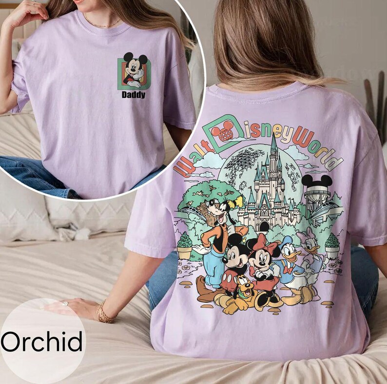 May include: Orchid-coloured t-shirt with a Mickey Mouse graphic and the word "Daddy" on the front. The back features a "Walt Disney World" design with Mickey, Minnie, Donald, Goofy, and Pluto. The shirt is a light purple colour.