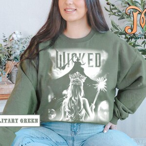 May include: Military green sweatshirt featuring a white graphic of a woman with a crown and a wand, with the word "WICKED" above her. The sweatshirt is a crew neck style.