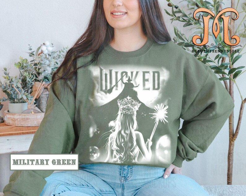 May include: Military green sweatshirt featuring a white graphic of a woman with a crown and a wand, with the word "WICKED" above her. The sweatshirt is a crew neck style.