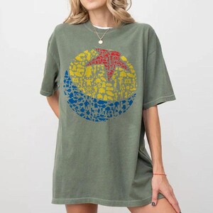 May include: Olive green t-shirt featuring a colorful graphic design. The design is a circle divided into three sections: a red star, a yellow middle, and a blue bottom, all filled with various character silhouettes.
