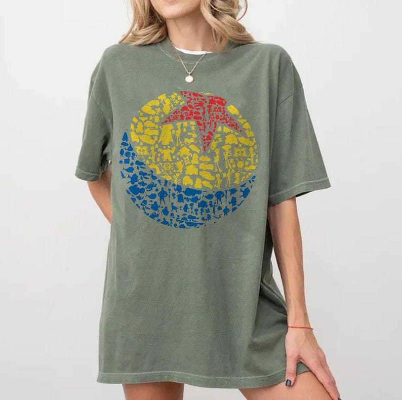 May include: Olive green t-shirt featuring a colorful graphic design. The design is a circle divided into three sections: a red star, a yellow middle, and a blue bottom, all filled with various character silhouettes.
