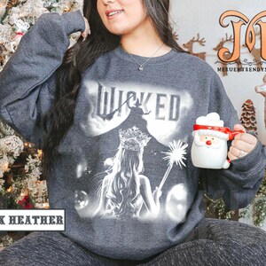 May include: Dark heather gray sweatshirt with a graphic of the word "WICKED" and a silhouetted witch and princess. The sweatshirt also features the text "DARK HEATHER". A Santa mug is held in the hand.