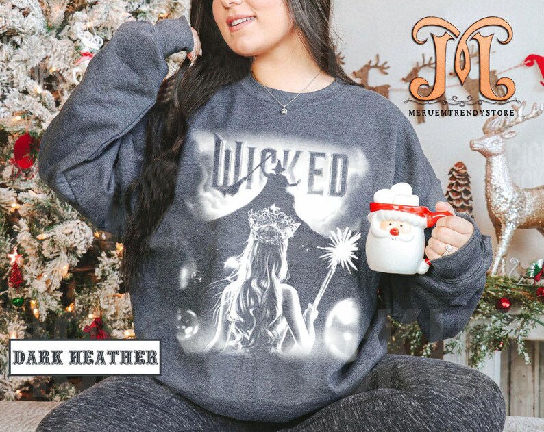 May include: Dark heather gray sweatshirt with a graphic of the word "WICKED" and a silhouetted witch and princess. The sweatshirt also features the text "DARK HEATHER". A Santa mug is held in the hand.