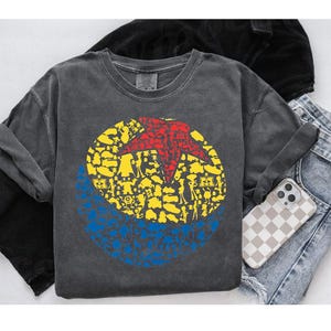 May include: A charcoal gray long-sleeve shirt featuring a circular graphic with a red star and yellow and blue shapes. The shirt is paired with denim jeans and a phone case with a checkered pattern.