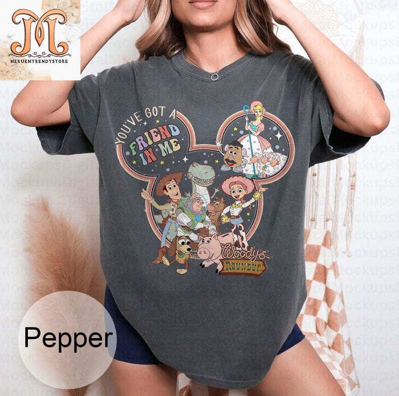 Puede incluir: Una camiseta gris oscuro con un gr&aacute;fico de cabeza de Mickey Mouse de colores con personajes de la franquicia Toy Story. El gr&aacute;fico incluye el texto "You've got a friend in me" y "Woody's Roundup".