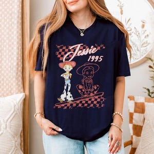 K&ouml;nnte beinhalten: Marineblaues T-Shirt mit einem Aufdruck von Jessie aus Toy Story, mit dem Text "Jessie 1995" in einem rosa-wei&szlig; karierten Design. Das Design enth&auml;lt zwei Illustrationen von Jessie in ihrem Cowboy-Outfit.