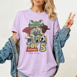 May include: A light purple t-shirt featuring a graphic from the Disney Pixar movie Toy Story 5. The design includes characters like Woody, Buzz Lightyear, and Jessie, with the movie title in a bold, colorful font. The shirt is a casual wear item.