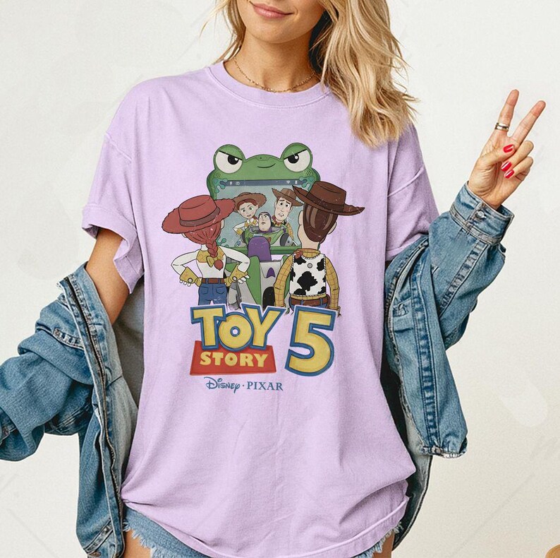 May include: A light purple t-shirt featuring a graphic from the Disney Pixar movie Toy Story 5. The design includes characters like Woody, Buzz Lightyear, and Jessie, with the movie title in a bold, colorful font. The shirt is a casual wear item.