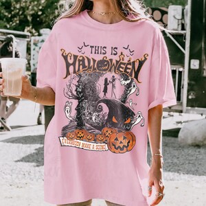 May include: A light pink t-shirt featuring a graphic print with the text "THIS IS HALLOWEEN" and "EVERYBODY MAKE A SCENE". The design includes silhouettes of two characters, ghosts, bats, a dark tree, and several orange jack-o'-lantern pumpkins.