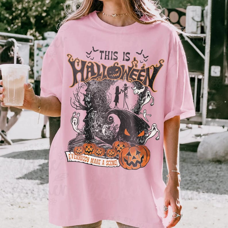 May include: A light pink t-shirt featuring a graphic print with the text "THIS IS HALLOWEEN" and "EVERYBODY MAKE A SCENE". The design includes silhouettes of two characters, ghosts, bats, a dark tree, and several orange jack-o'-lantern pumpkins.