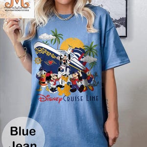 May include: Blue jean t-shirt featuring a colorful graphic of Disney characters on a cruise ship, with palm trees and a sun. The text "Disney Cruise Line" is displayed below the graphic. The shirt has short sleeves.