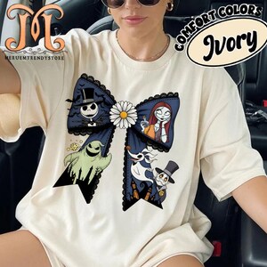 Peut inclure: T-shirt ivoire avec un grand motif de n&oelig;ud orn&eacute; de personnages de L'&Eacute;trange No&euml;l de Monsieur Jack. Le n&oelig;ud est bleu marine avec une bordure en dentelle noire et une marguerite blanche. Le t-shirt porte le texte "COMFORT COLORS" et "Ivory".