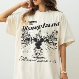 以下が含まれることがあります： クリーム色のTシャツ。ミッキーマウスがディズニーランドの街並みを背景にした白黒のグラフィックが特徴です。「CALIFORNIA .st. 1955 Disneyland」と「The happiest place on earth」の文字がプリントされています。
