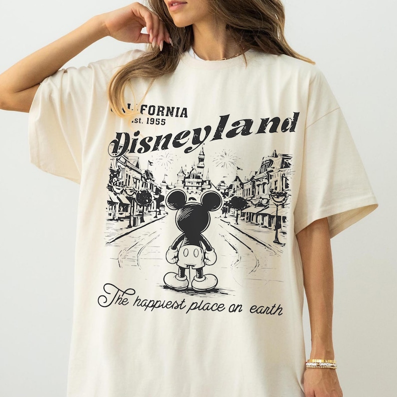 以下が含まれることがあります： クリーム色のTシャツ。ミッキーマウスがディズニーランドの街並みを背景にした白黒のグラフィックが特徴です。「CALIFORNIA .st. 1955 Disneyland」と「The happiest place on earth」の文字がプリントされています。