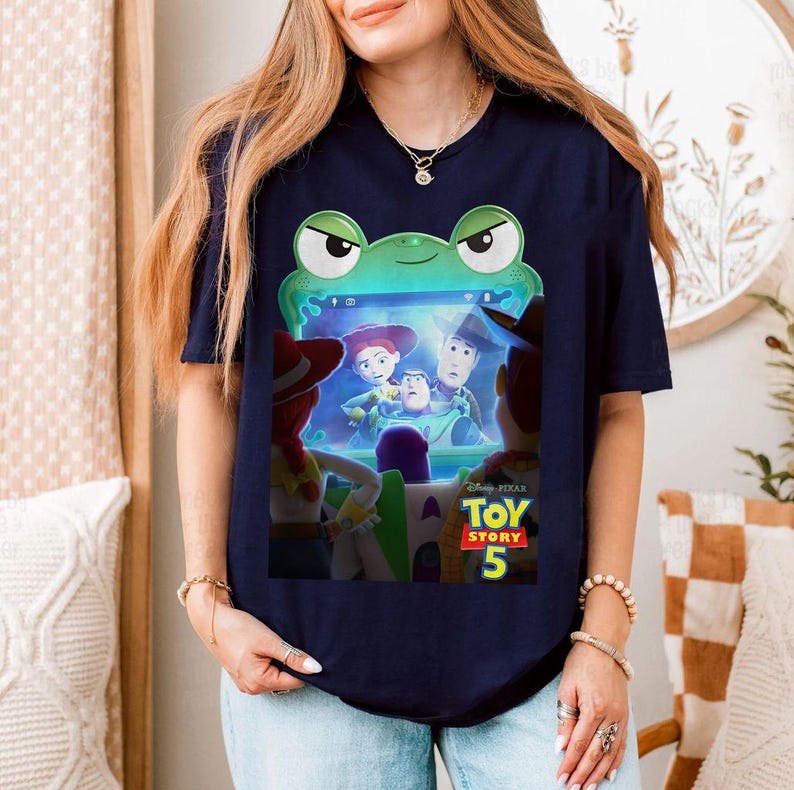 May include: Dark blue t-shirt with a vibrant graphic from the film Toy Story 5. The design showcases characters such as Woody and Buzz Lightyear, set within a green frog-themed frame. The top is a short-sleeved crew neck.