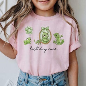 Peut inclure: T-shirt rose clair avec des caméléons verts de dessin animé et l'inscription "best day ever". Un caméléon tient des ballons, un autre est encadré dans un ovale doré et un troisième est sur une branche. Le t-shirt est porté par un enfant.