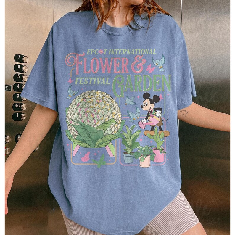May include: A light blue t-shirt featuring the text "EPCOT INTERNATIONAL FLOWER & GARDEN FESTIVAL". The graphic includes a geometric sphere, Mickey Mouse watering plants, and various flowers and butterflies.
