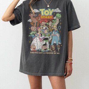 Puede incluir: Camiseta gris oscuro con un gr&aacute;fico colorido de los personajes de Toy Story. El dise&ntilde;o incluye el t&iacute;tulo de la pel&iacute;cula y la frase "You've Got a Friend in Me". La camiseta tiene un aspecto vintage, ligeramente descolorido.