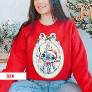 May include: A red sweatshirt featuring a cartoon character in a Santa hat and a bow tie, set within an ornate frame. The frame includes a castle and holly accents. The word "RED" is displayed in a white box at the bottom.