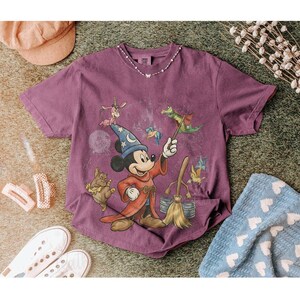 K&ouml;nnte beinhalten: Ein lila T-Shirt mit einer Grafik von Mickey Mouse als Zauberer, umgeben von Zeichentrickfiguren. Das Shirt hat einen Rundhalsausschnitt und kurze &Auml;rmel. Das Bild befindet sich auf einer strukturierten Oberfl&auml;che mit anderen Gegenst&auml;nden.