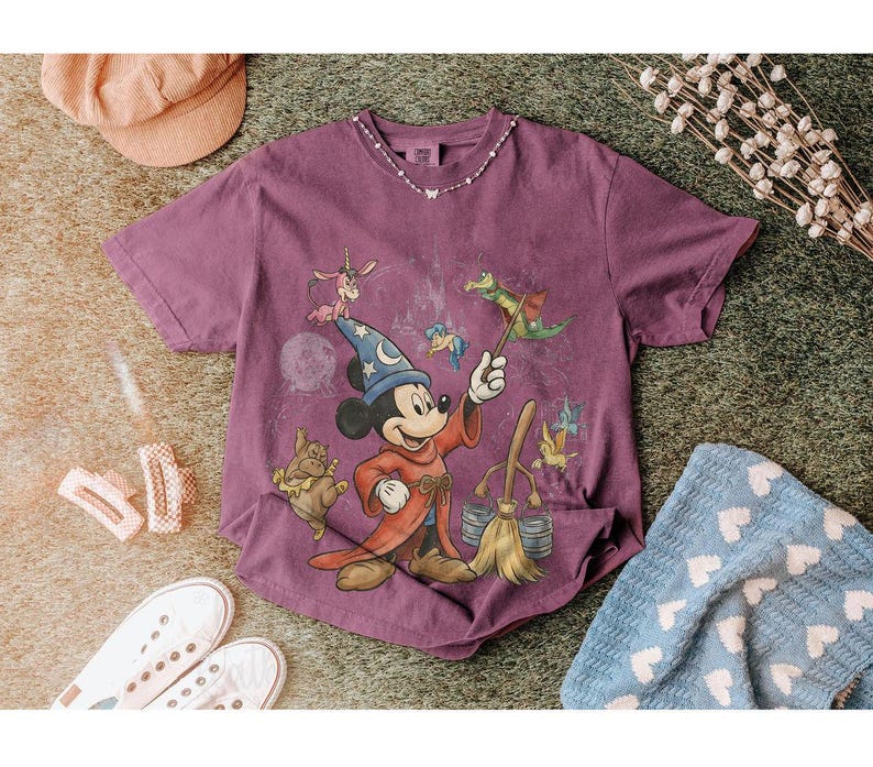 K&ouml;nnte beinhalten: Ein lila T-Shirt mit einer Grafik von Mickey Mouse als Zauberer, umgeben von Zeichentrickfiguren. Das Shirt hat einen Rundhalsausschnitt und kurze &Auml;rmel. Das Bild befindet sich auf einer strukturierten Oberfl&auml;che mit anderen Gegenst&auml;nden.