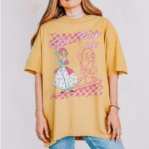 K&ouml;nnte beinhalten: Senfgelbes T-Shirt mit einer Grafik von Bo Peep aus Toy Story. Das Design umfasst zwei Illustrationen von Bo Peep, den Text "Bo Peep 1995" und ein rosa-wei&szlig;es Karomuster. Das Shirt hat kurze &Auml;rmel und eine lockere Passform.